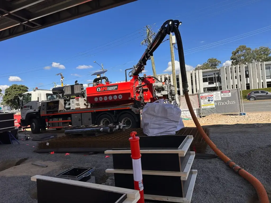  A red and white vacuum excavation truck from APU Excavations & Rentals is onsite, using its boom arm and suction hose to remove material into a large white bulka bag. The jobsite is active with safety markers, reinforcing mesh, and concrete formwork in place, with commercial buildings and signage in the background under a clear blue sky.
